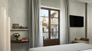 A bedroom with a large bed, a TV mounted on the wall, a window with curtains, and a shelf with decorative items. - NH Collection Palacio de Córdoba (Córdoba)