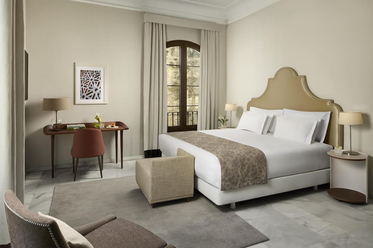 Una habitación de hotel con cama, escritorio, silla, ventana y lámpara.