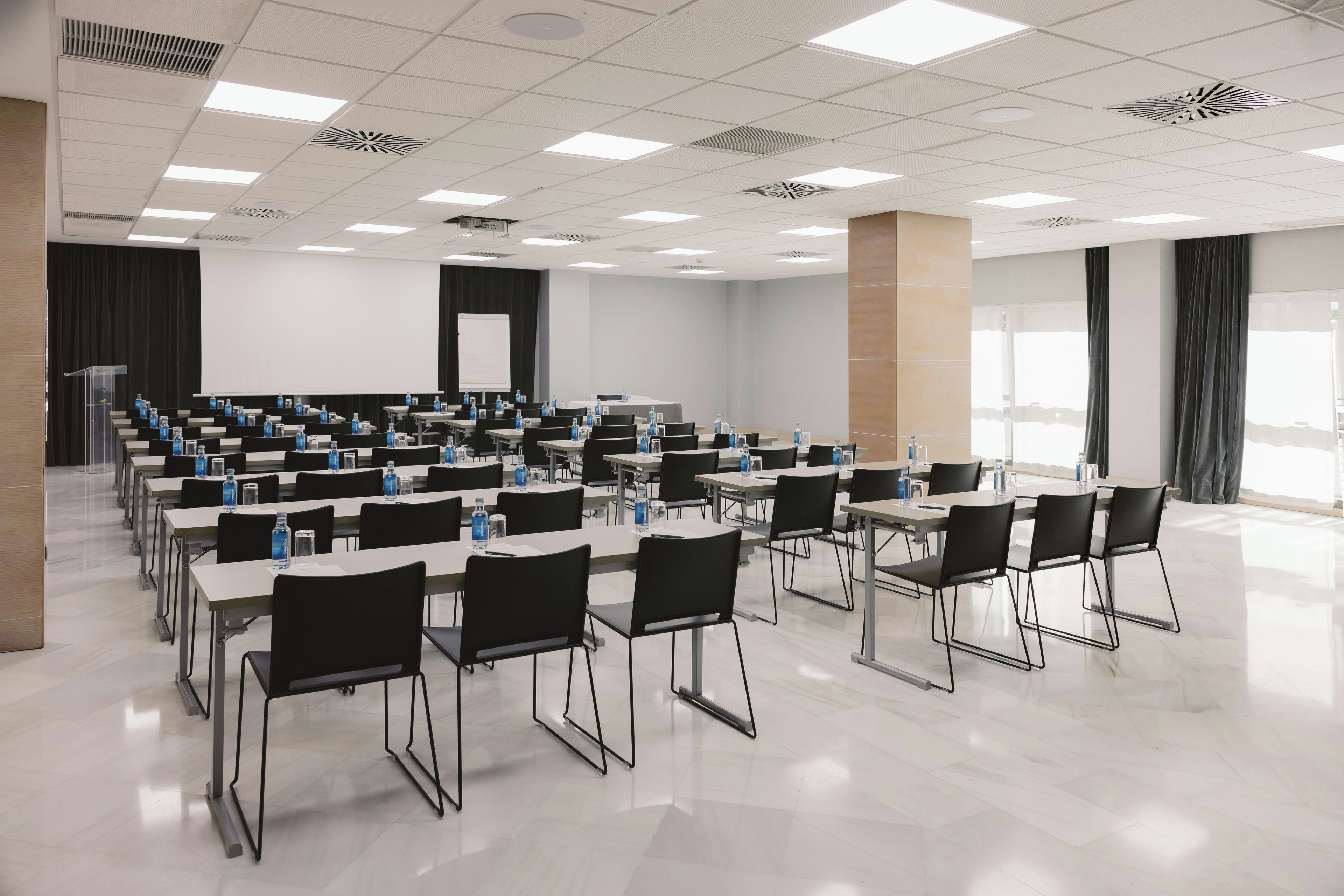 A conference room with rows of tables and chairs, equipped with water bottles, and a projection screen at the front.