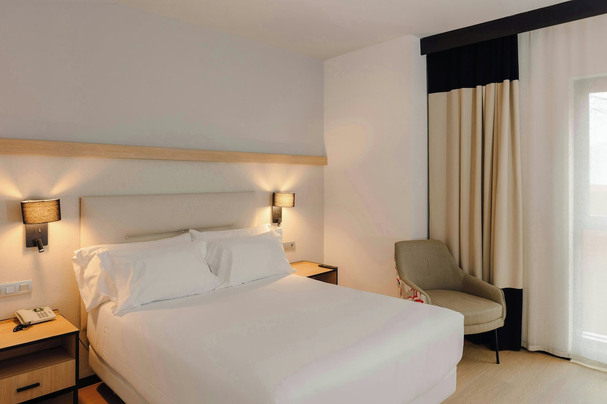 Una habitación de hotel con cama, lámparas de noche, una silla y una ventana con cortinas.