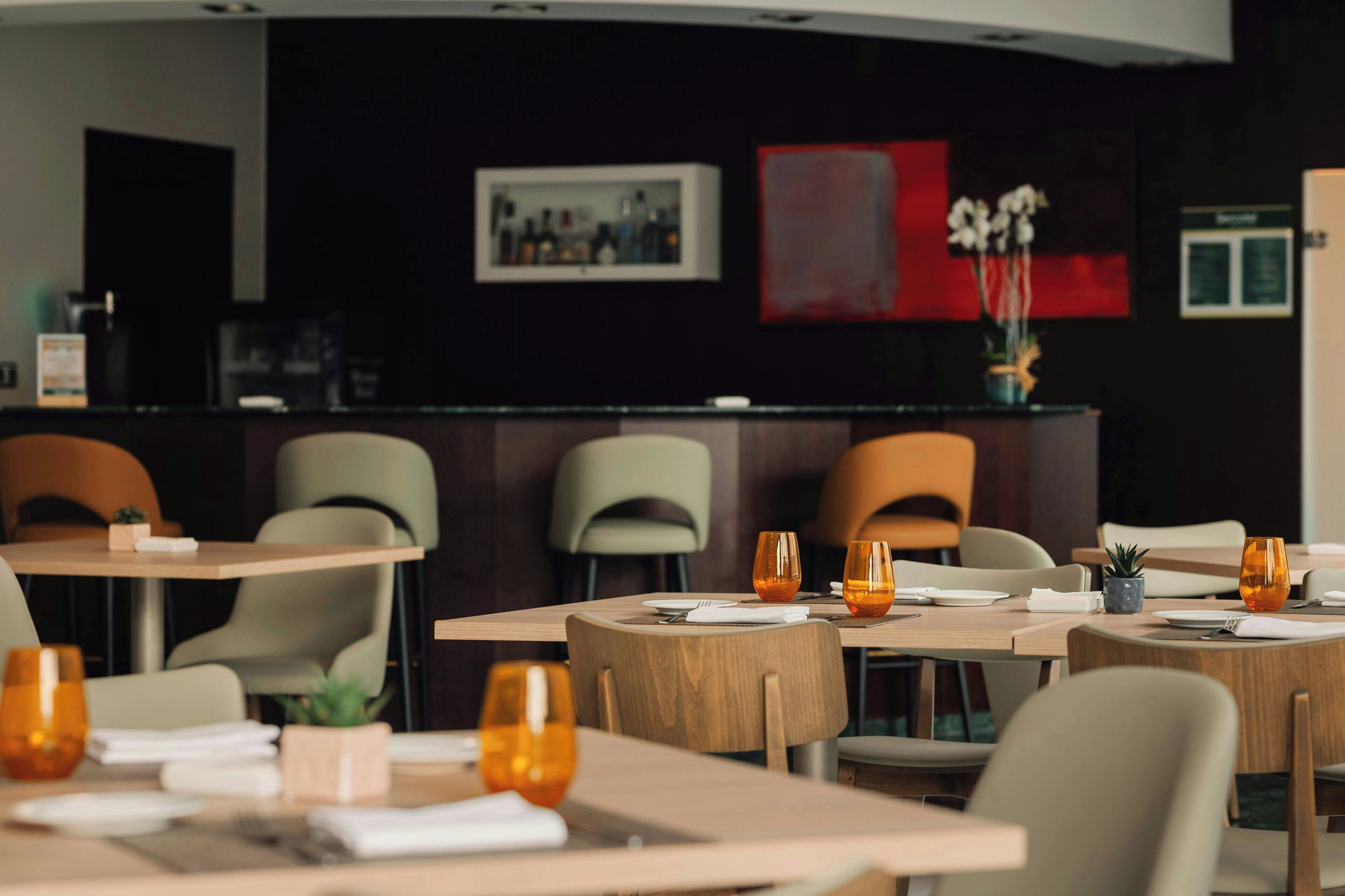 A restaurant with wooden tables and chairs, some with orange glasses. There are framed pictures on the wall and a plant on the counter.