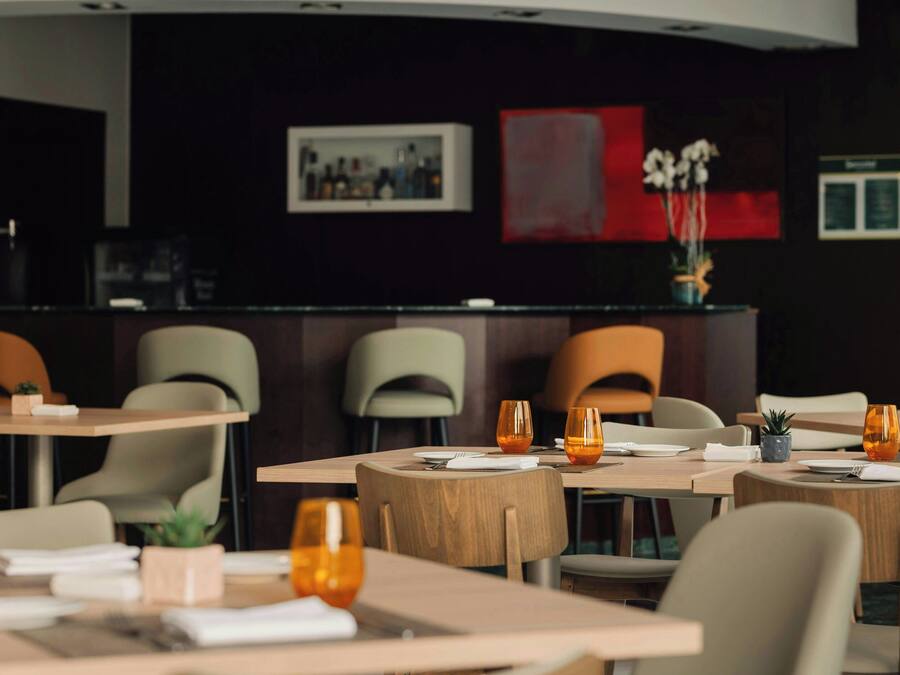 A restaurant with wooden tables and chairs, some with orange glasses. There are framed pictures on the wall and a plant on the counter.