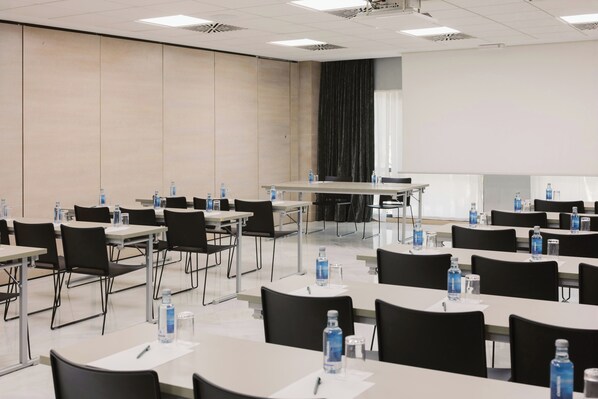Un salón de conferencias con varias mesas y sillas, equipado con botellas de agua y bolígrafos, y una pantalla de proyector al frente.
