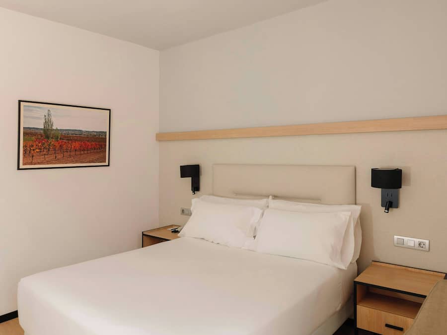 A hotel room with a bed, bedside tables, a wall-mounted TV, and a framed picture of a vineyard.
