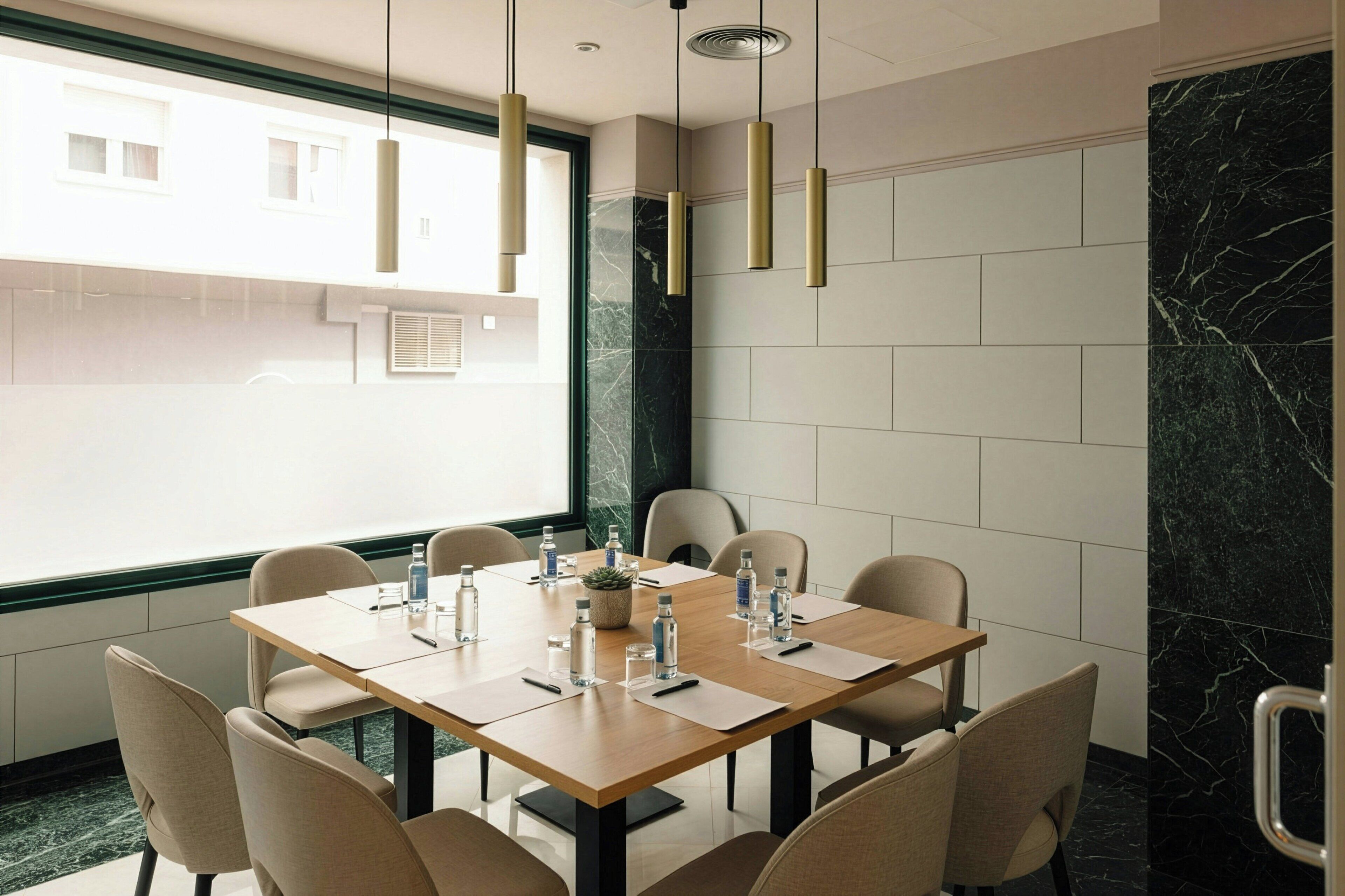 Un salón de conferencias con una mesa de madera, sillas color beige, botellas de agua y una planta en maceta.