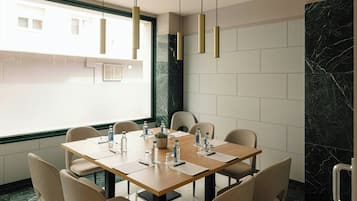 Un salón de conferencias con una mesa de madera, sillas color beige, botellas de agua y una planta en maceta.