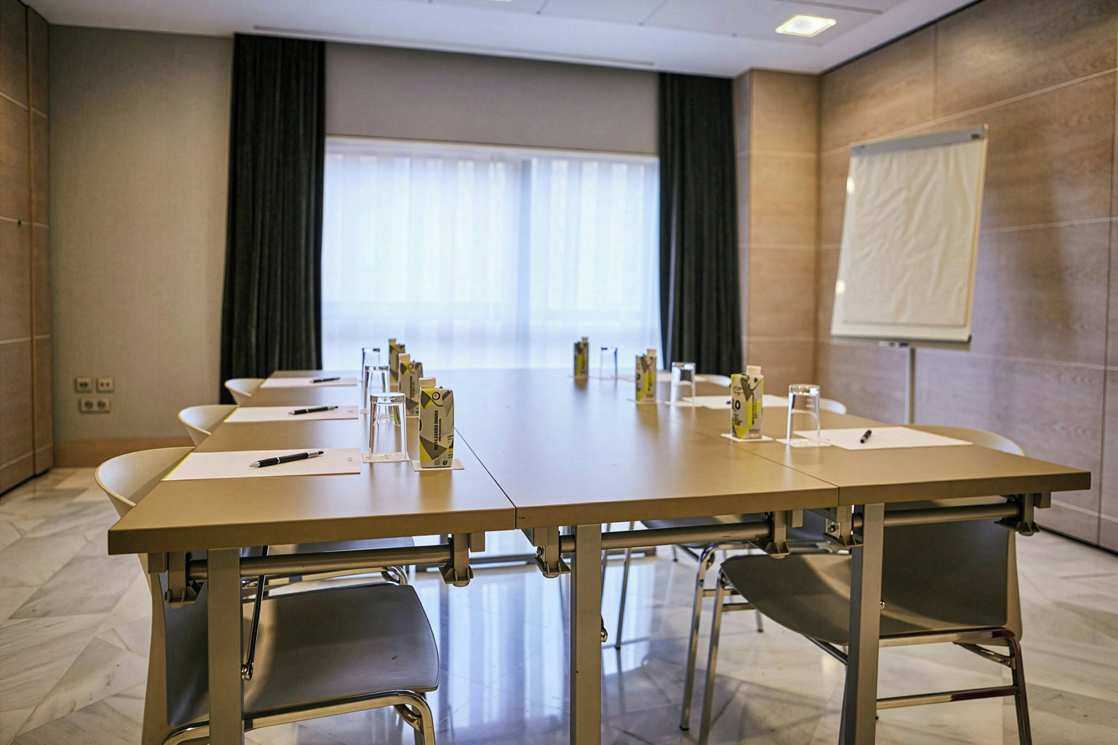 Un salón de conferencias con una larga mesa, sillas, una pizarra y una ventana con cortinas.