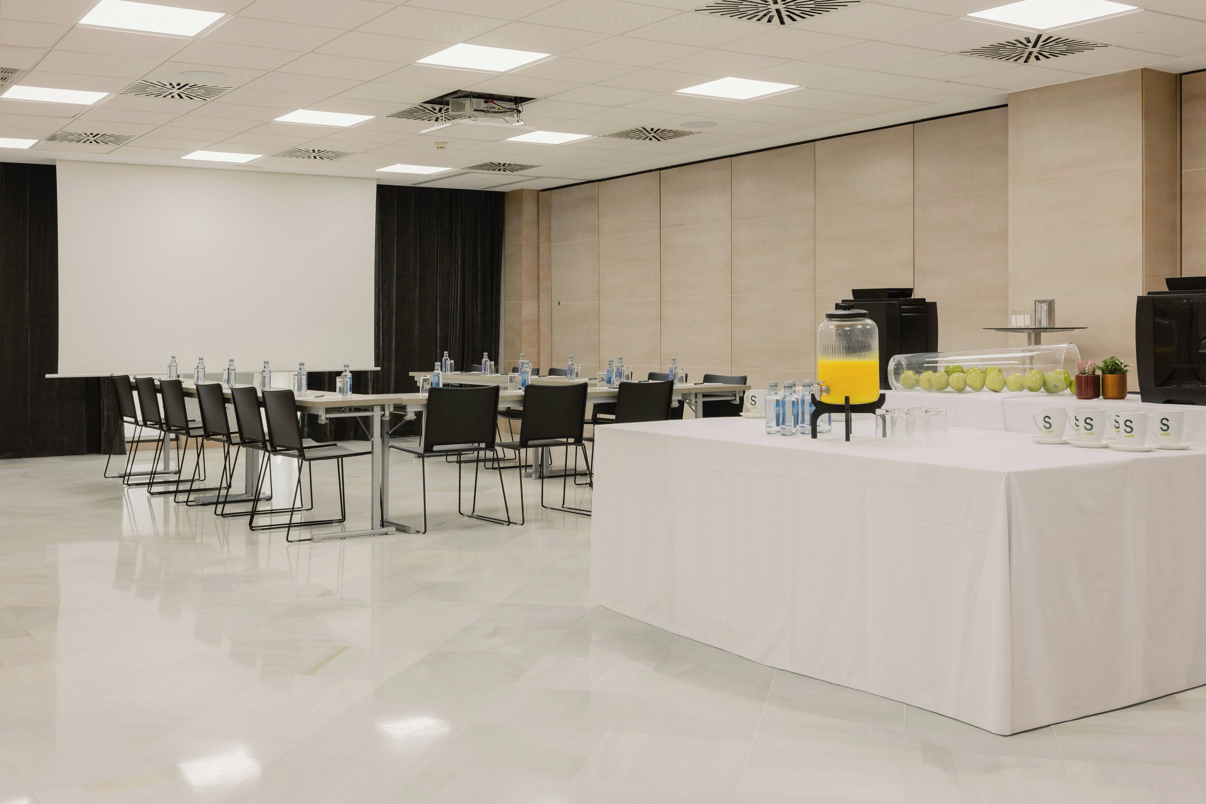 Une salle de conférence équipée d’une longue table, de chaises, d’un projecteur et d’un coin rafraîchissements avec un présentoir de fruits.