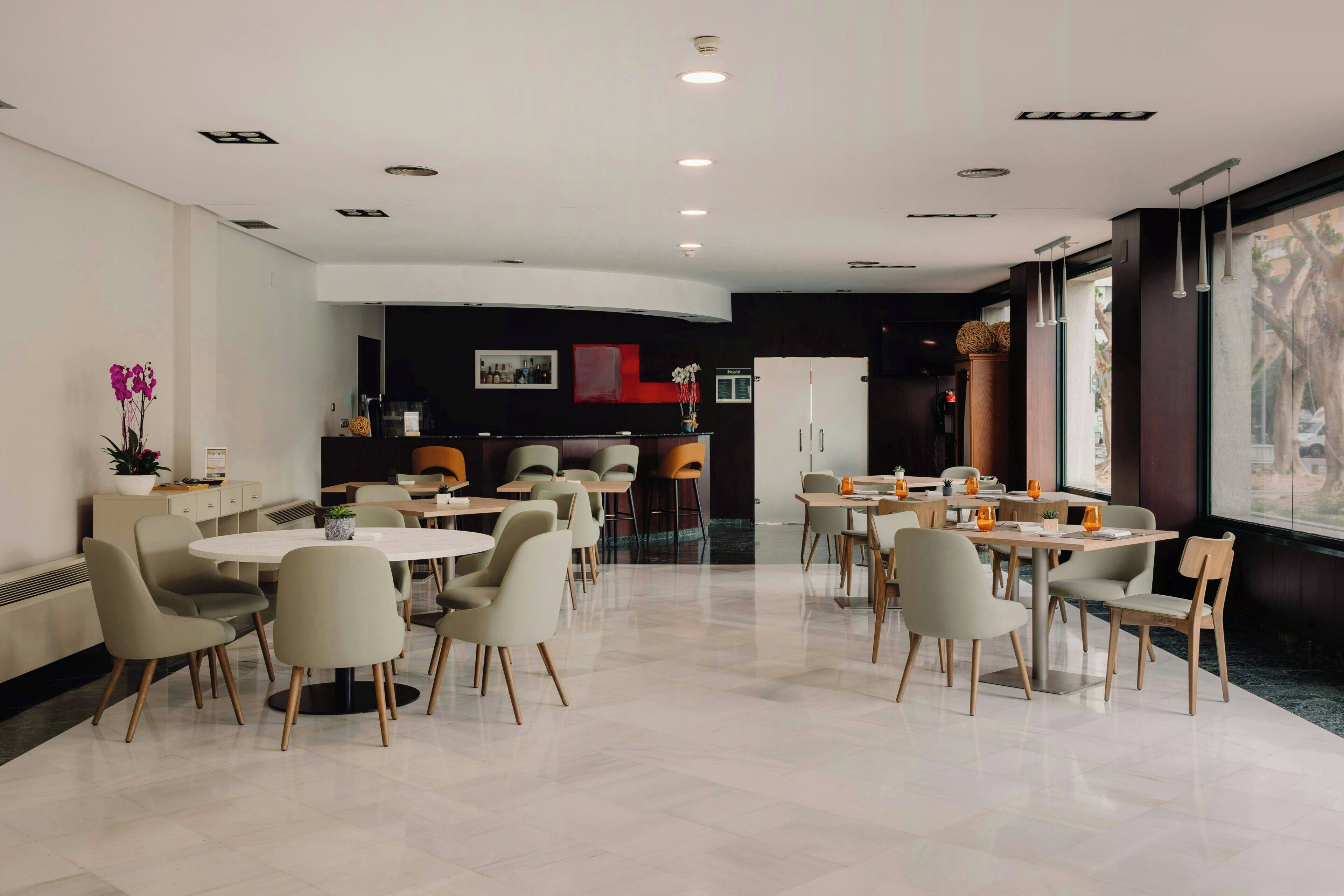 A modern hotel lobby with round tables and chairs, a reception desk, and large windows.