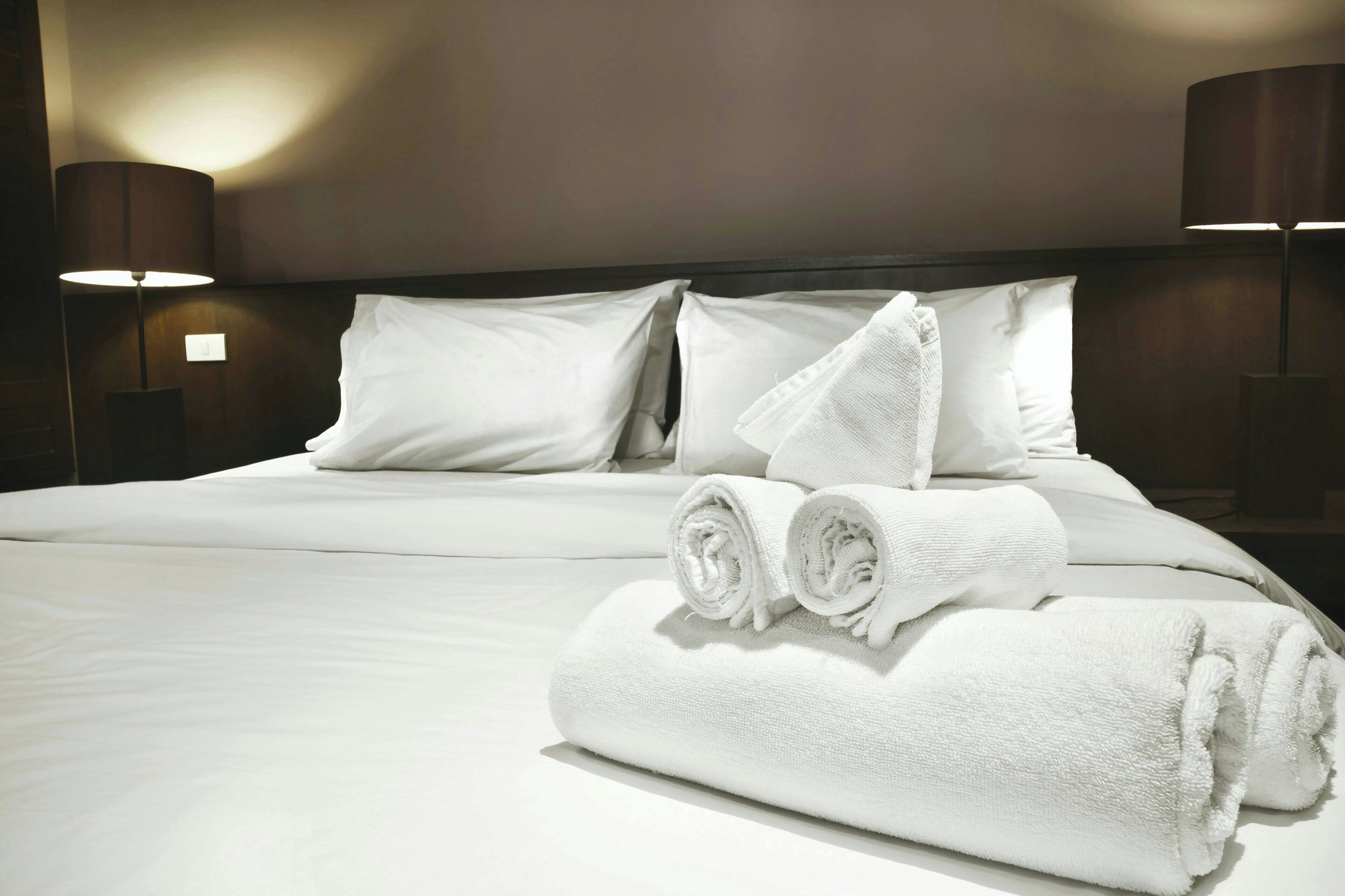 A neatly made bed with white linens and towels, two bedside lamps, and a plain wall.