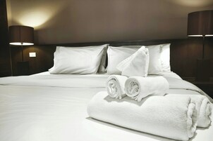 A neatly made bed with white linens and towels, two bedside lamps, and a plain wall.