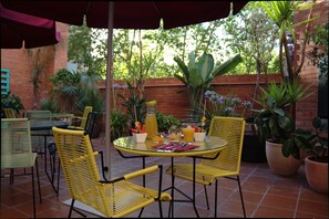 An outdoor patio with yellow wicker chairs and a glass table, surrounded by potted plants and a brick wall. - Chic & Basic Velvet (Barcelona)