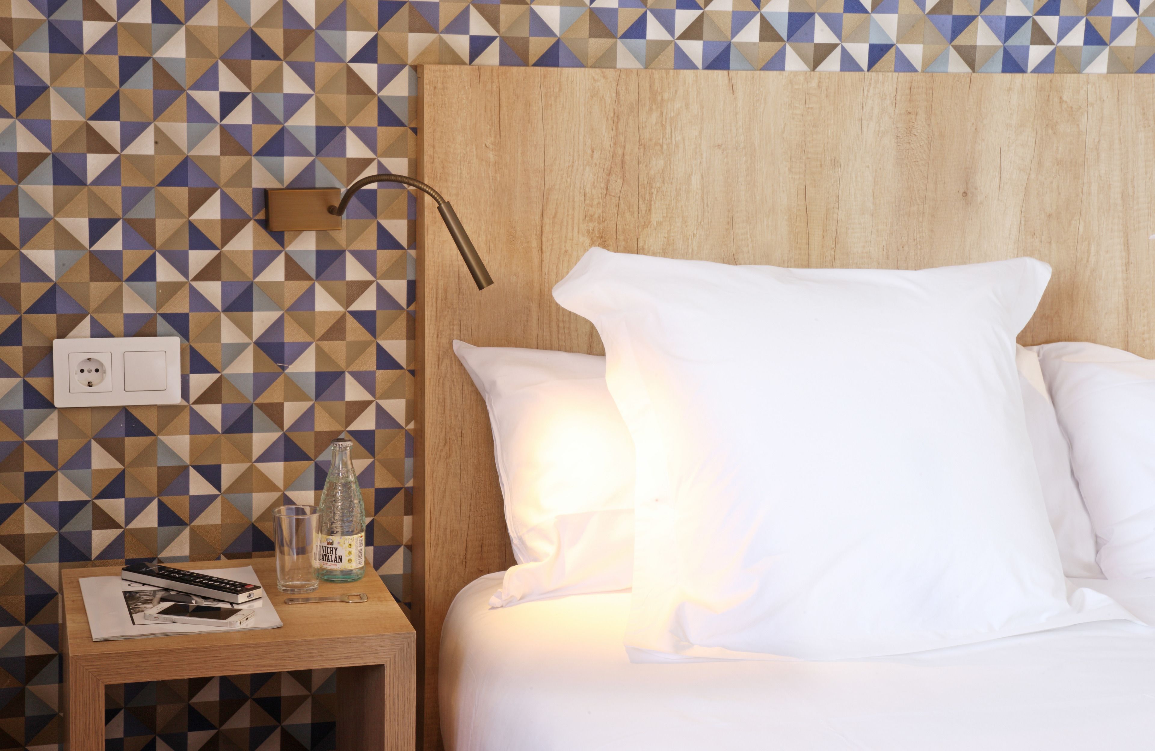 A neatly made bed with white linens, a bedside table with a remote control, a glass, and a bottle of water.