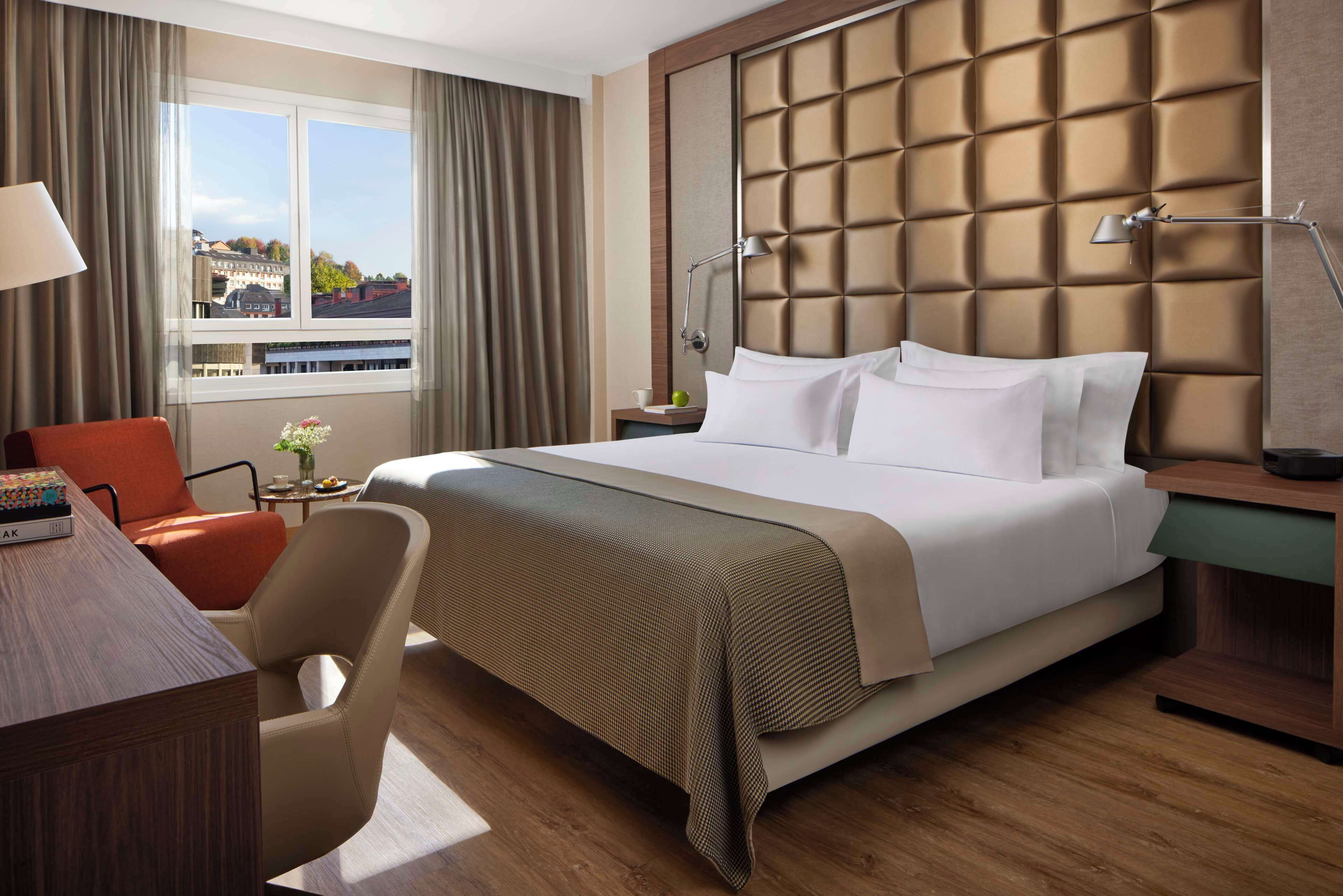 Habitación de hotel con una cama grande, un escritorio, una silla, una mesita de noche y una ventana con vistas a edificios.