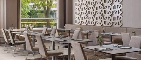 A modern dining area with multiple tables and chairs, large windows with a view of greenery, and a decorative wall panel.
