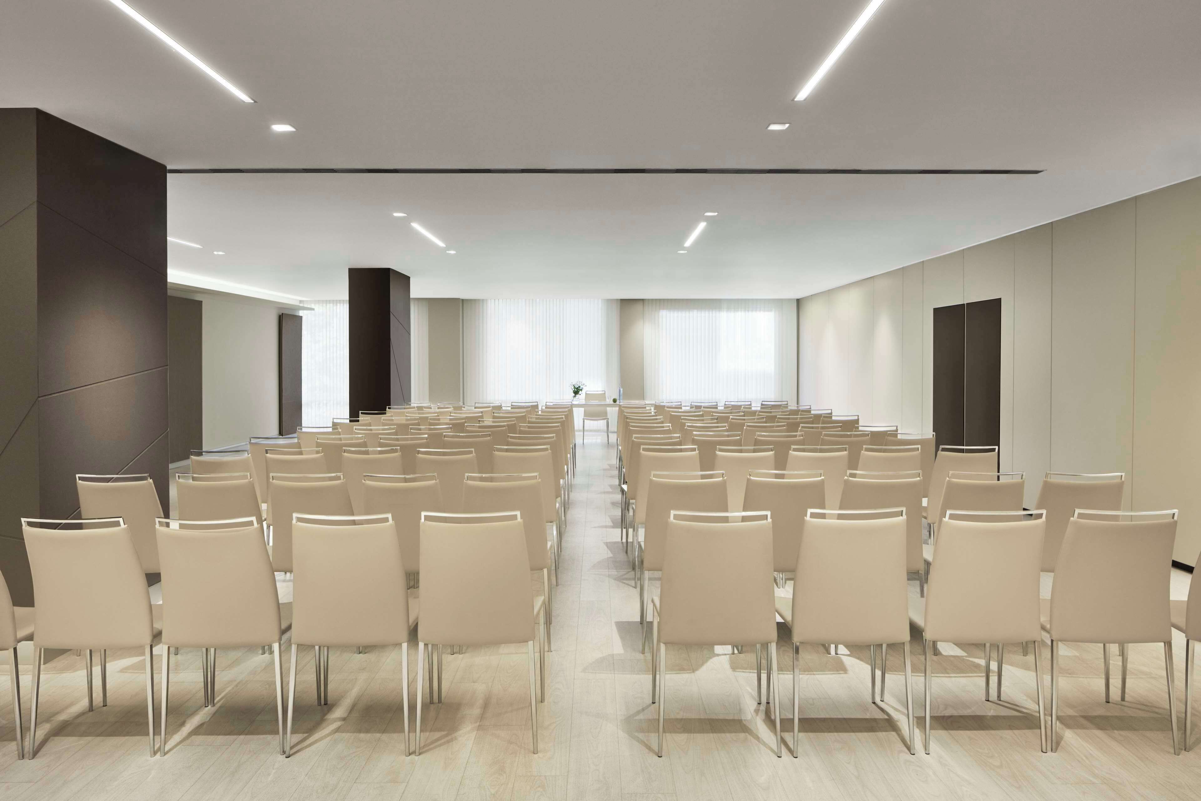 Une salle de conférence avec des rangées de chaises disposées face à une scène, équipée de luminaires au plafond et de grandes fenêtres.
