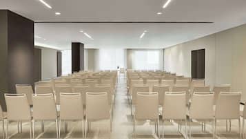 Une salle de conférence avec des rangées de chaises disposées face à une scène, équipée de luminaires au plafond et de grandes fenêtres.