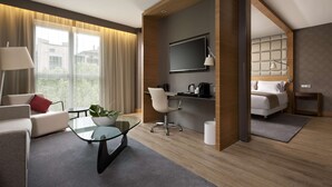 Ein modernes Hotelzimmer mit Sofa, Glastisch, Schreibtisch mit Stuhl, Bett, Fernseher und Blick ins Freie.