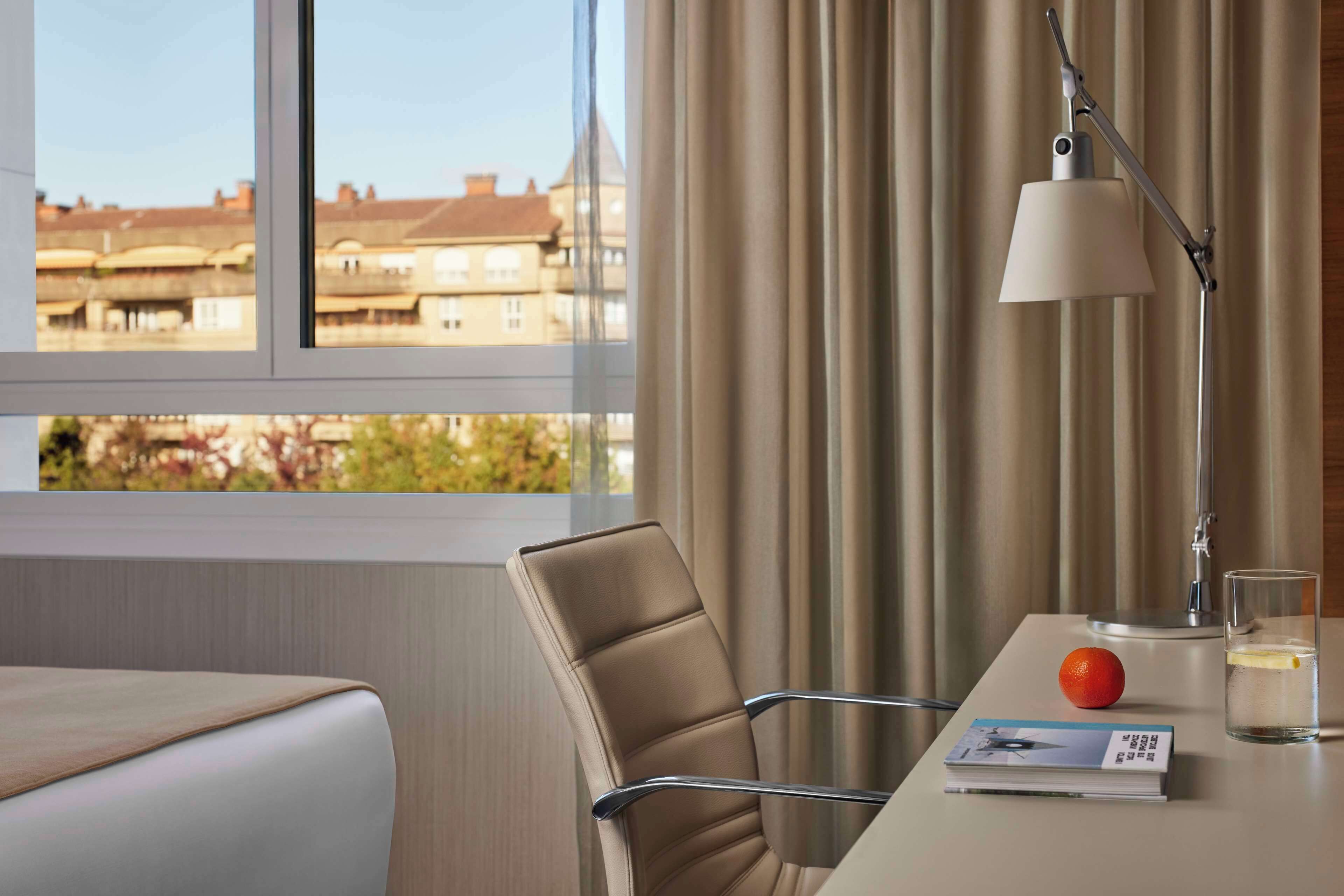A hotel room with a view of buildings through the window, a table with a book, an orange, and a glass of water.