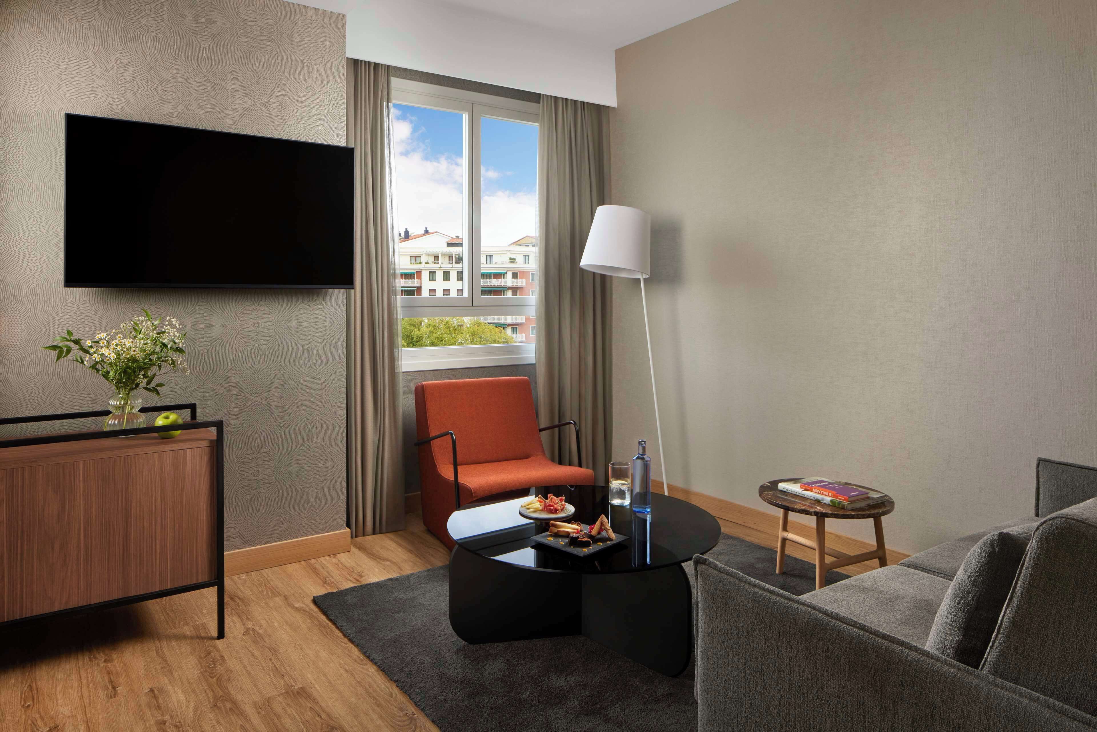 A modern hotel room with a flat-screen TV mounted on the wall, a comfortable orange chair, a grey sofa, a small round coffee table, and a side table with a lamp and a book.