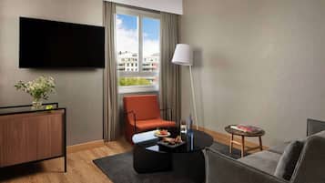 A modern hotel room with a flat-screen TV mounted on the wall, a comfortable orange chair, a grey sofa, a small round coffee table, and a side table with a lamp and a book.