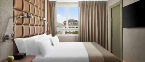 A hotel room with a large bed, a flat-screen TV, a bedside table with a lamp, and a view of buildings through the window.