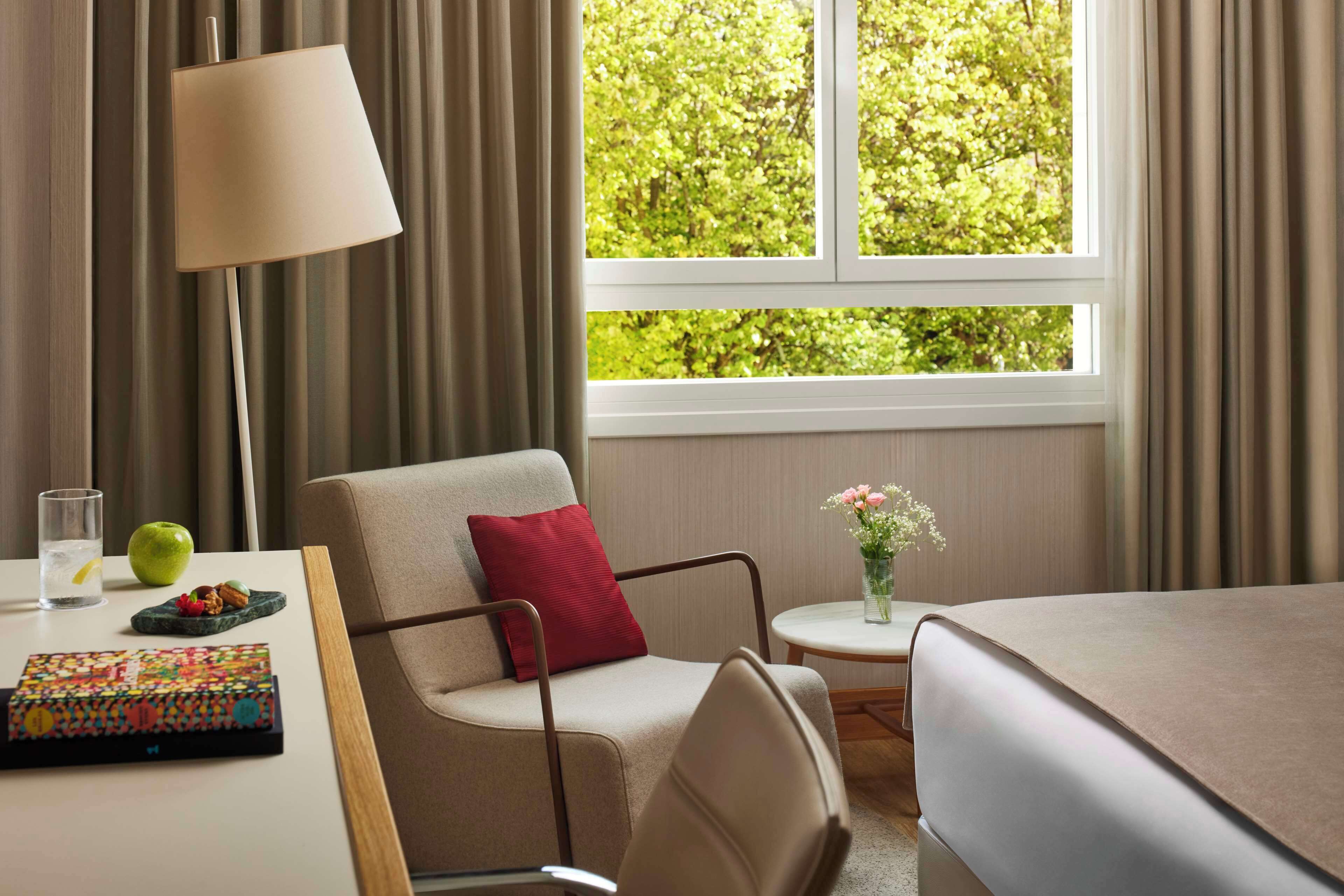 Una camera d'albergo con un letto, una sedia, un tavolo con una lampada, un vaso con fiori e una finestra con tende.
