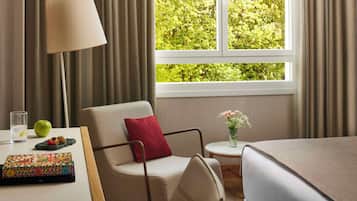 Una camera d'albergo con un letto, una sedia, un tavolo con una lampada, un vaso con fiori e una finestra con tende.