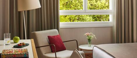 A hotel room with a bed, a chair, a table with a lamp, a vase with flowers, and a window with curtains.