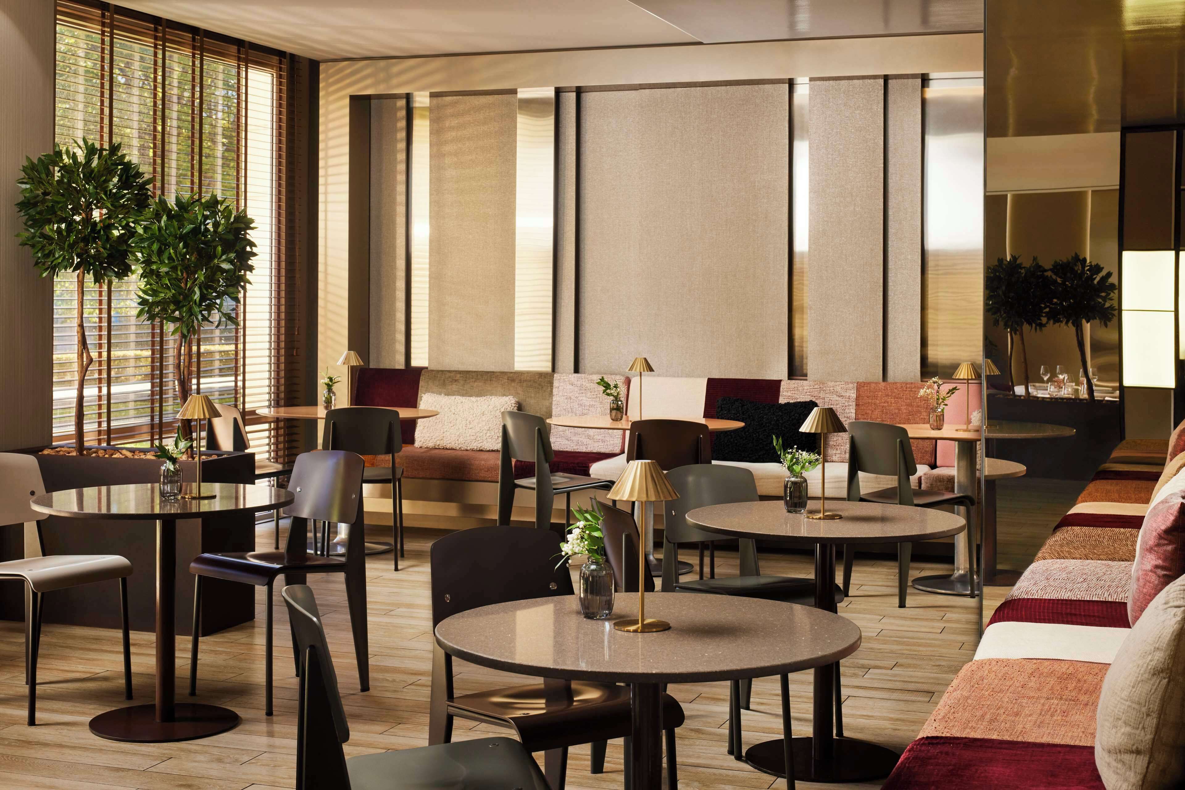 Un moderno vestíbulo de hotel con mesas redondas, asientos cómodos y amplias ventanas con persianas.