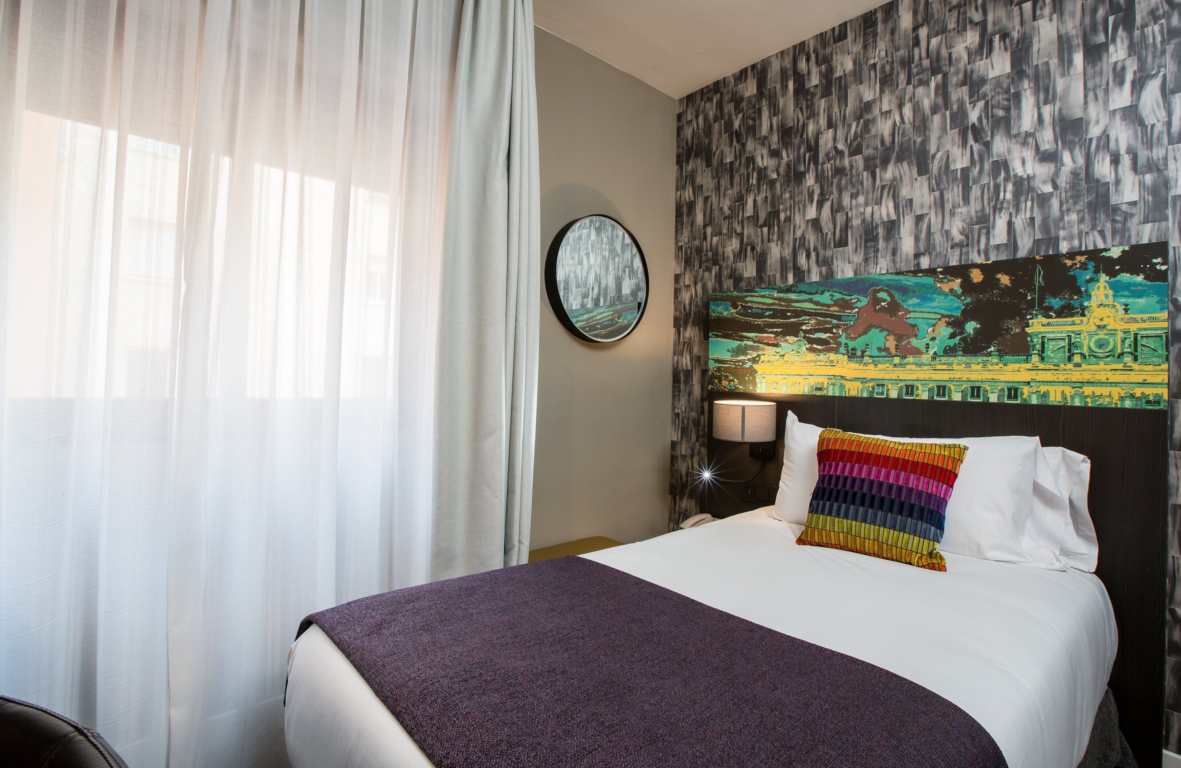 A hotel room with a large bed, a round wall clock, a colorful painting, and a window with sheer curtains.