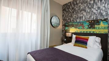 A hotel room with a large bed, a round wall clock, a colorful painting, and a window with sheer curtains.