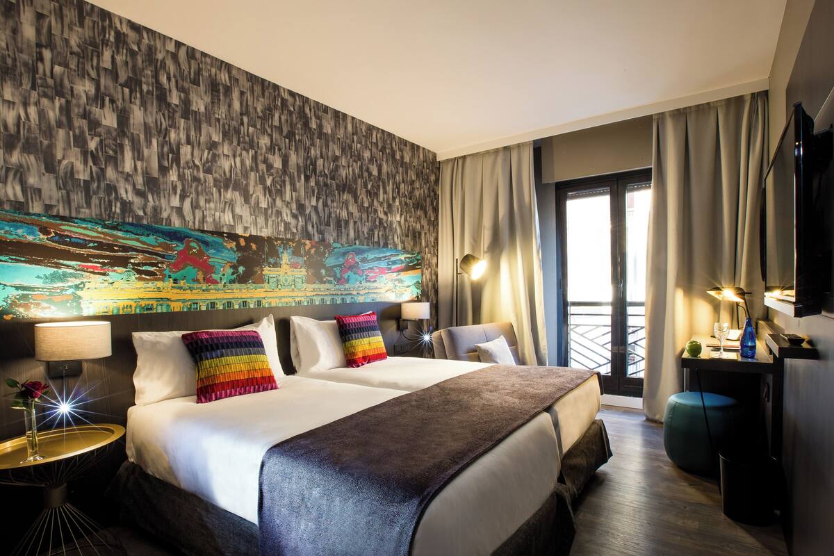 A hotel room with a large bed, two bedside tables with lamps, a TV, and a view of the city through the window.