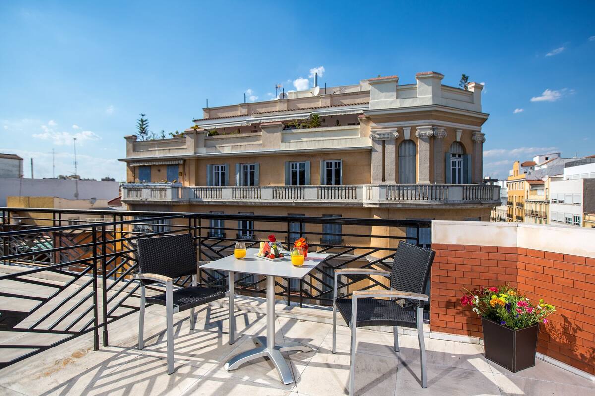 A rooftop terrace with outdoor seating, a flower pot, and a building with balconies in the background.