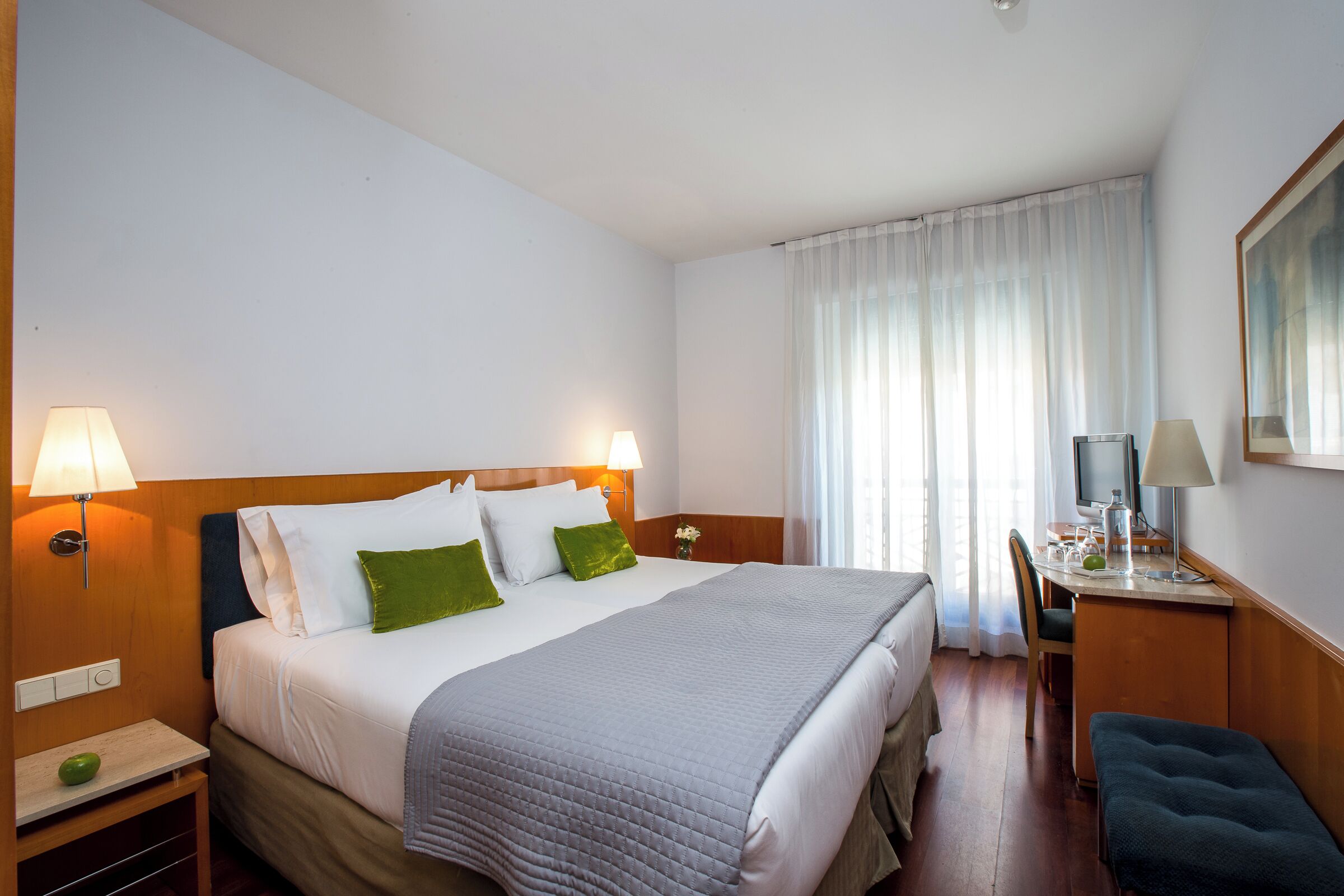 A hotel room with a bed, bedside lamps, a desk with a chair, a small table with a lamp, and a window with curtains.