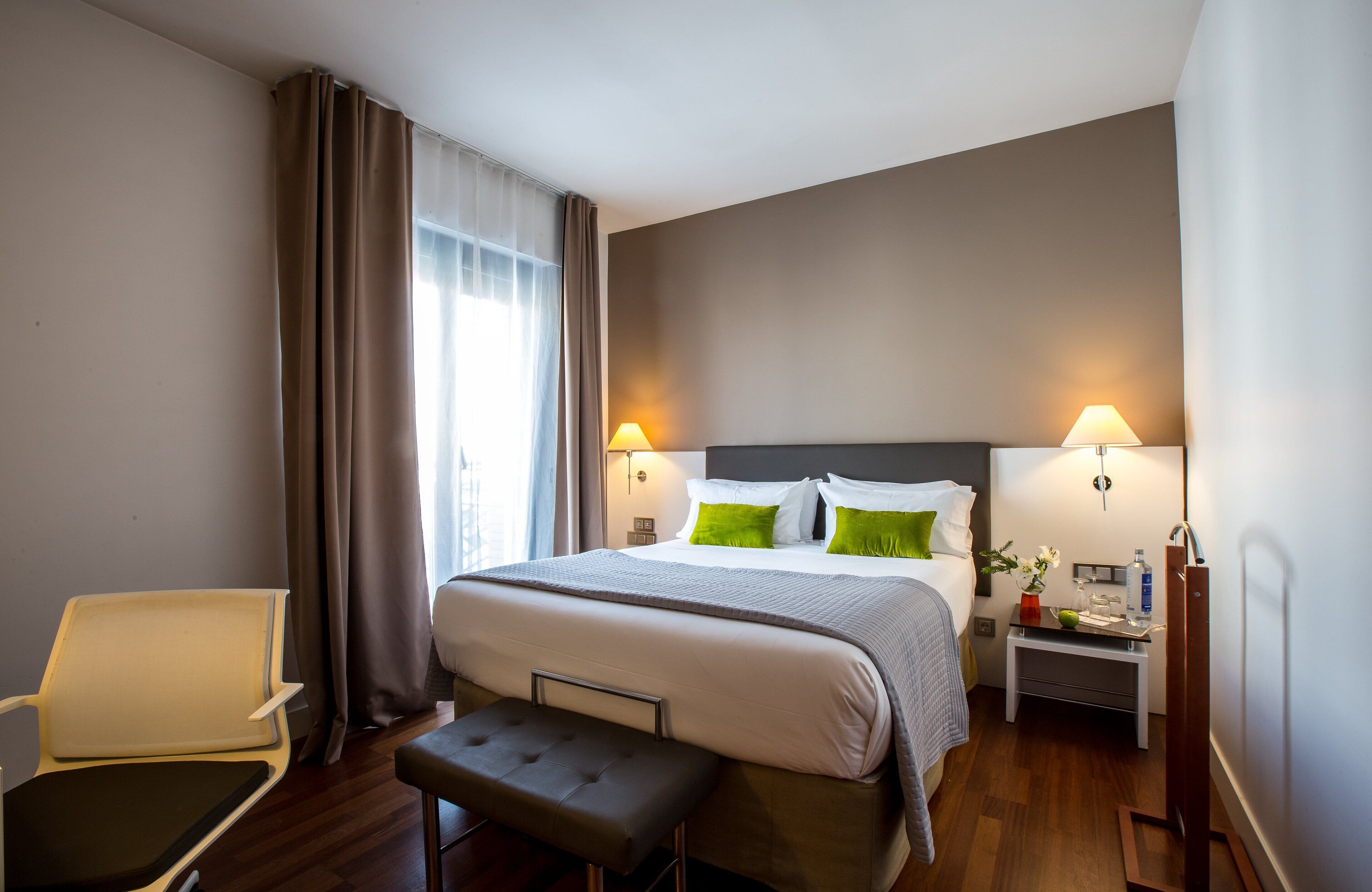 Une chambre d’hôtel avec un lit, des lampes de chevet, une petite table avec un vase, un banc et une chaise.