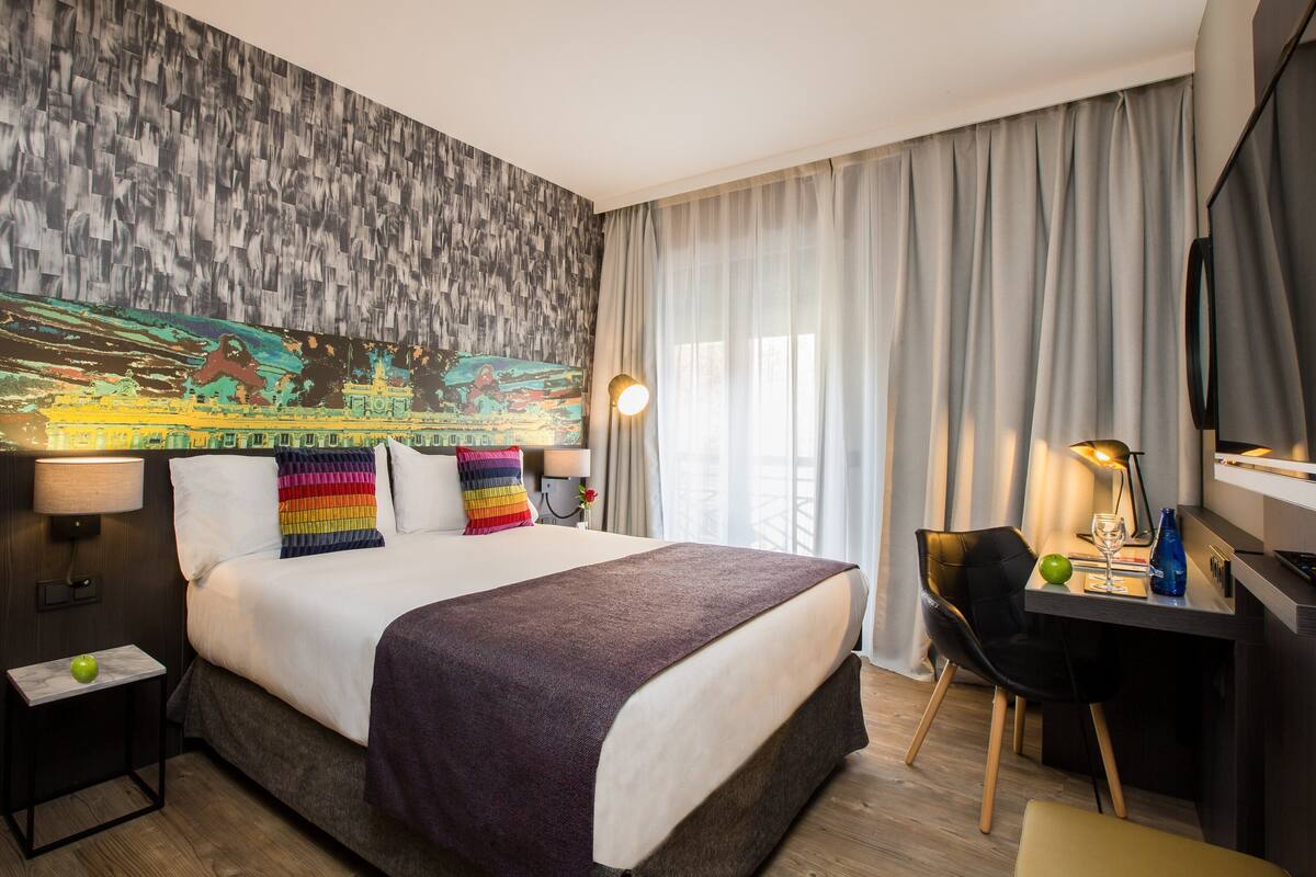 A hotel room with a large bed, a desk with a chair, a lamp, and a television.