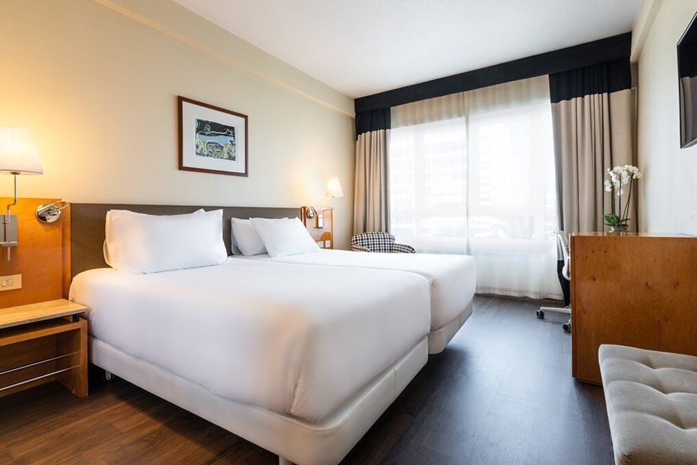 A hotel room with two beds, a desk, a chair, and a large window with curtains.