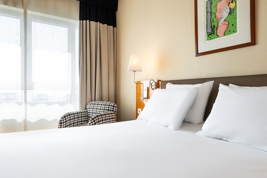 Ein Hotelzimmer mit einem großen Bett, einem Sessel, einer Lampe und einem fensterbepflanzten Fenster.