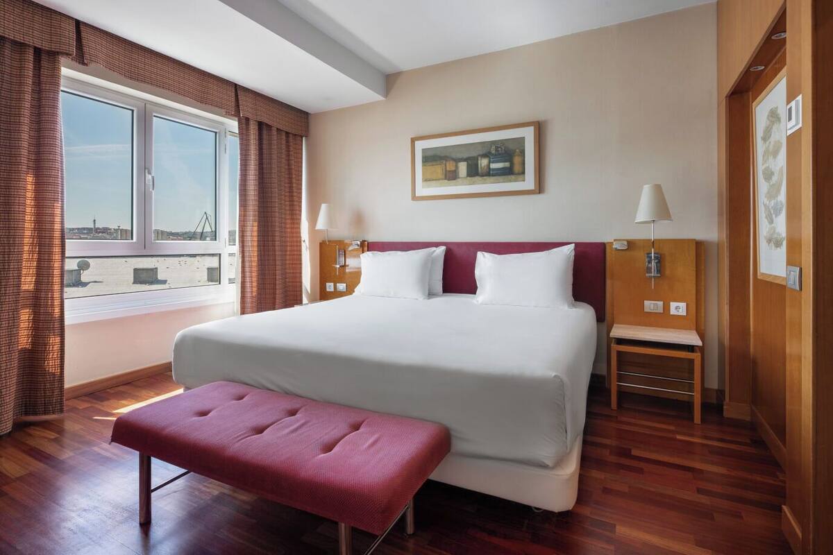 Ein Hotelzimmer mit einem großen Bett, einer Sitzbank, einem Fenster mit Vorhängen und einem Bild an der Wand.