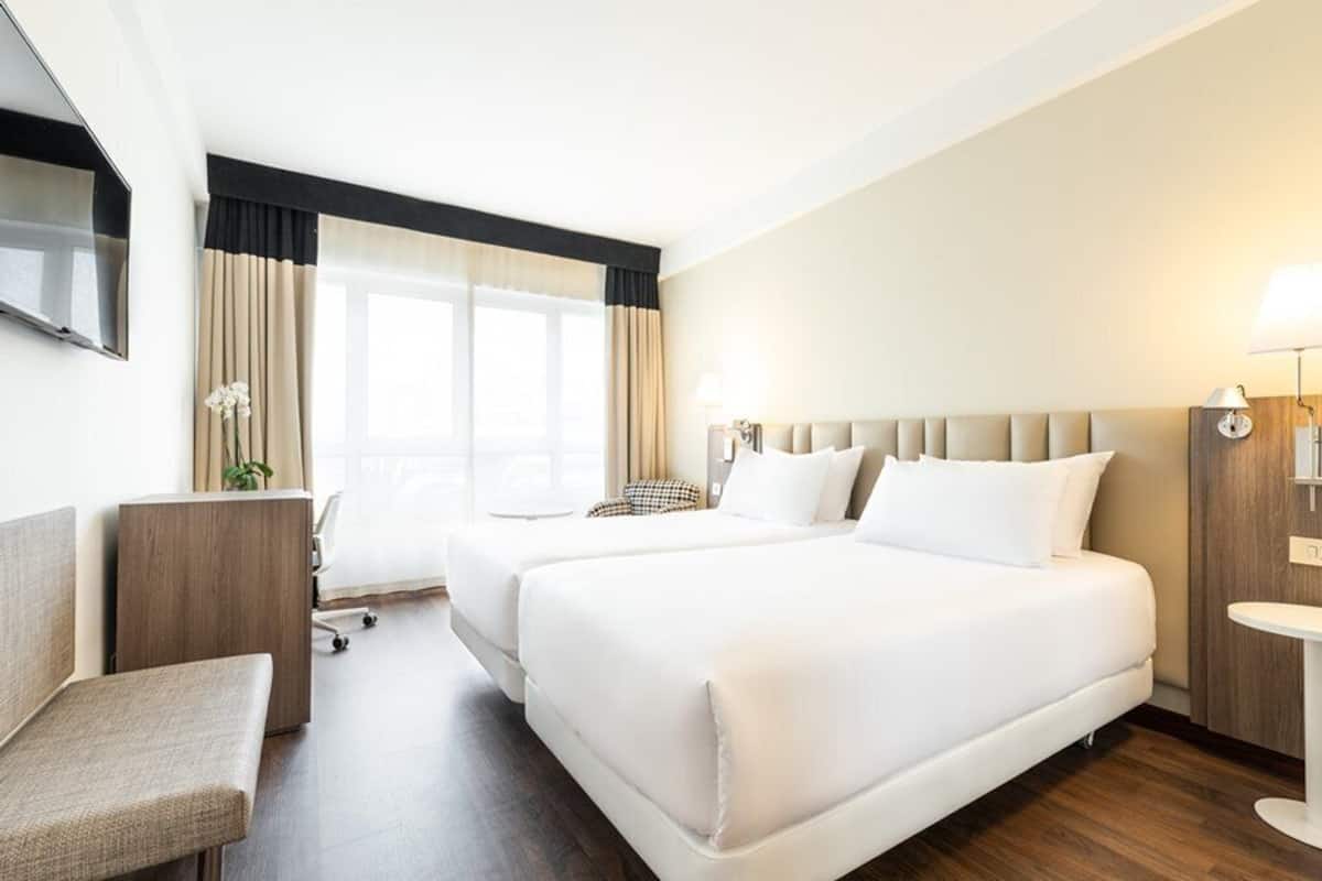 Ein Hotelzimmer mit zwei Betten, einem Schreibtisch, einem Sessel, einem Fernseher und einem Fenster mit Vorhängen.