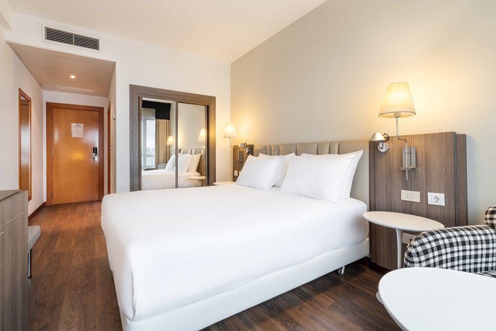 A hotel room with a large bed, a bedside table, a mirror, and a wall-mounted lamp.