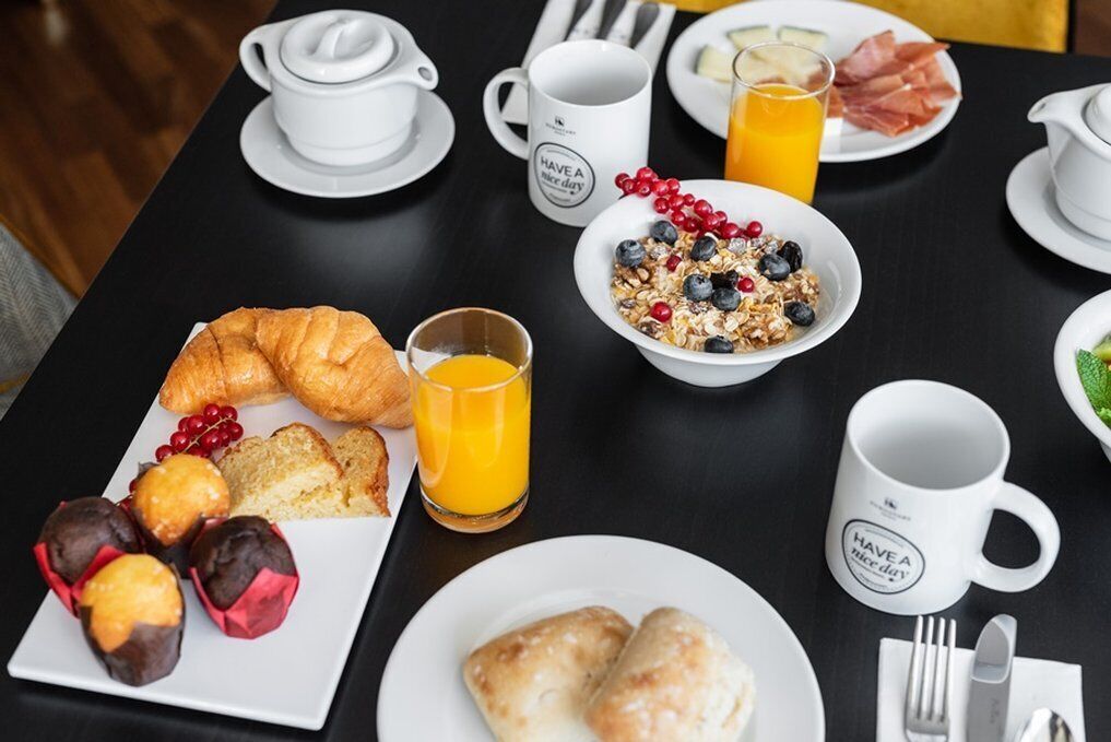 Una mesa de desayuno con croissants, pasteles, un tazón de cereales con bayas, jugo de naranja y una taza con una frase motivadora.