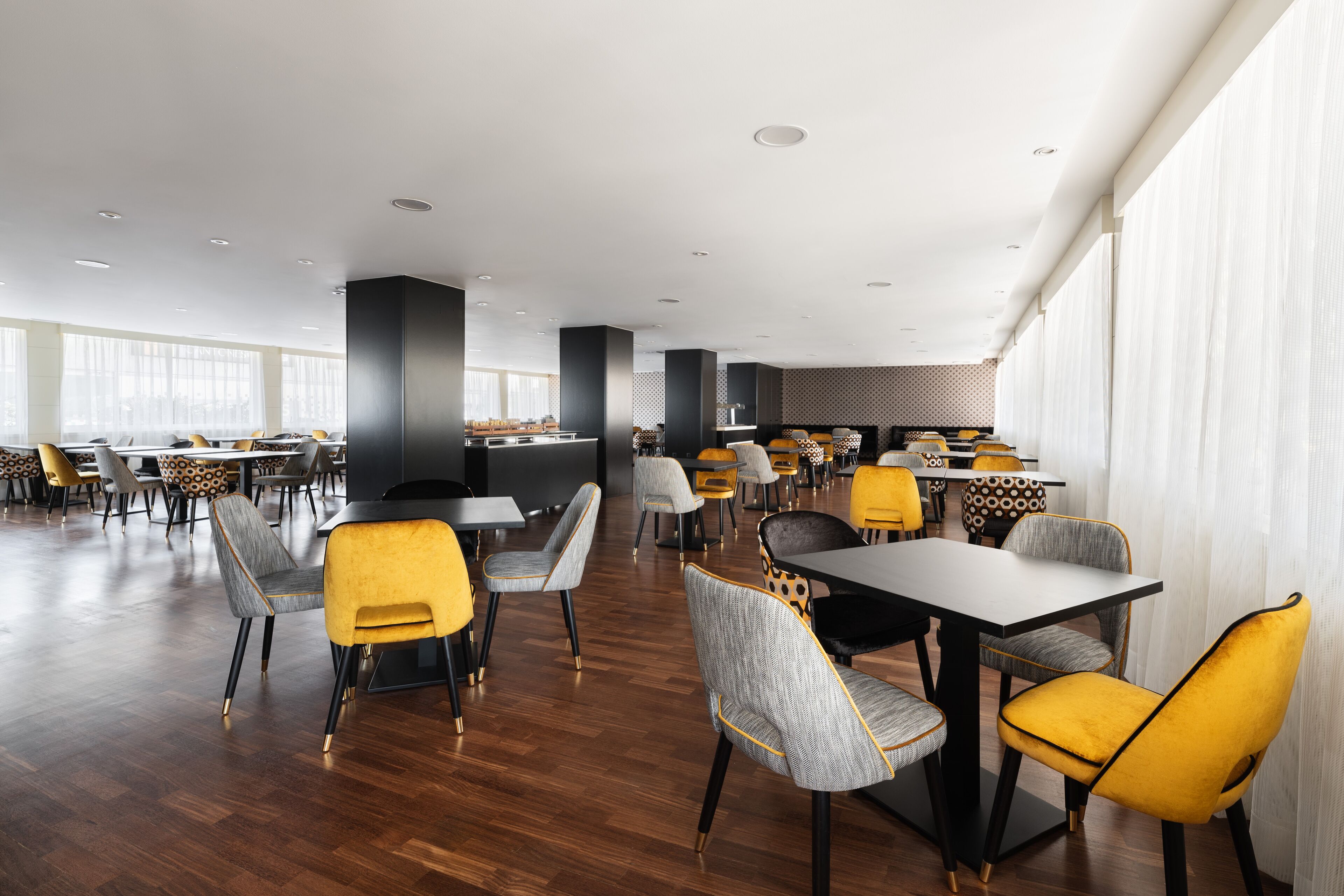 A spacious dining area with multiple tables and chairs, featuring a mix of yellow and grey upholstered chairs.