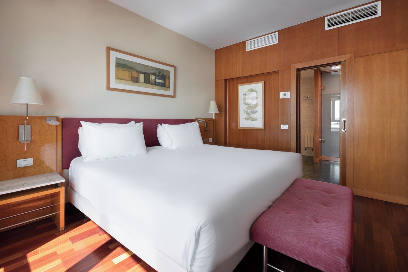 A hotel room with a large bed, wooden headboard, two bedside tables with lamps, a bench at the foot of the bed, and a door leading to another room.
