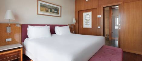 A hotel room with a large bed, wooden headboard, two bedside tables with lamps, a bench at the foot of the bed, and a door leading to another room.