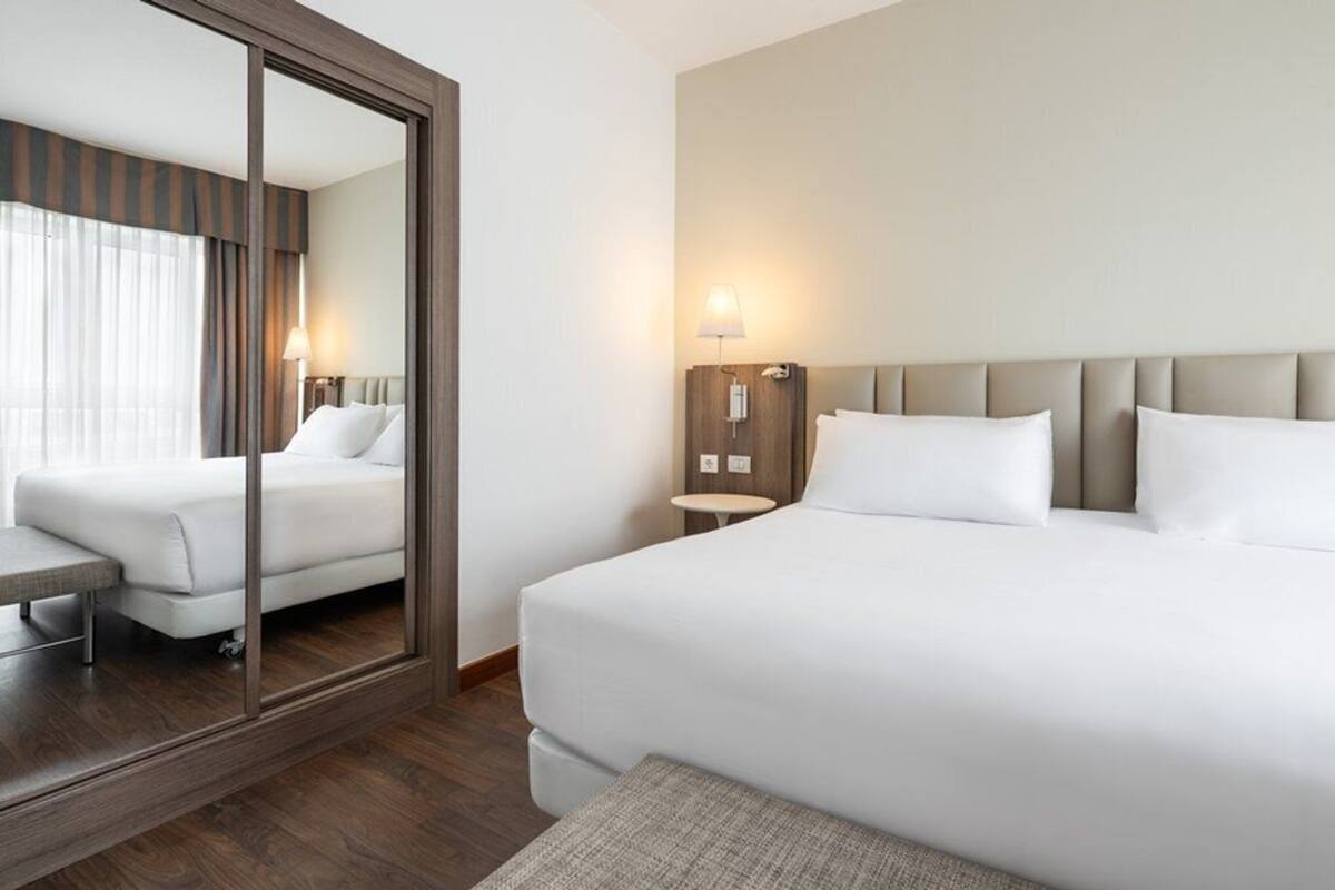 Ein Hotelzimmer mit einem großen Bett, einem Nachttisch mit Lampe, einem Spiegel und Blick auf ein weiteres Zimmer.