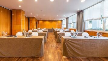 A conference room with multiple tables, each covered with a brown tablecloth, equipped with chairs and water glasses. The room has wooden flooring and large windows with curtains.