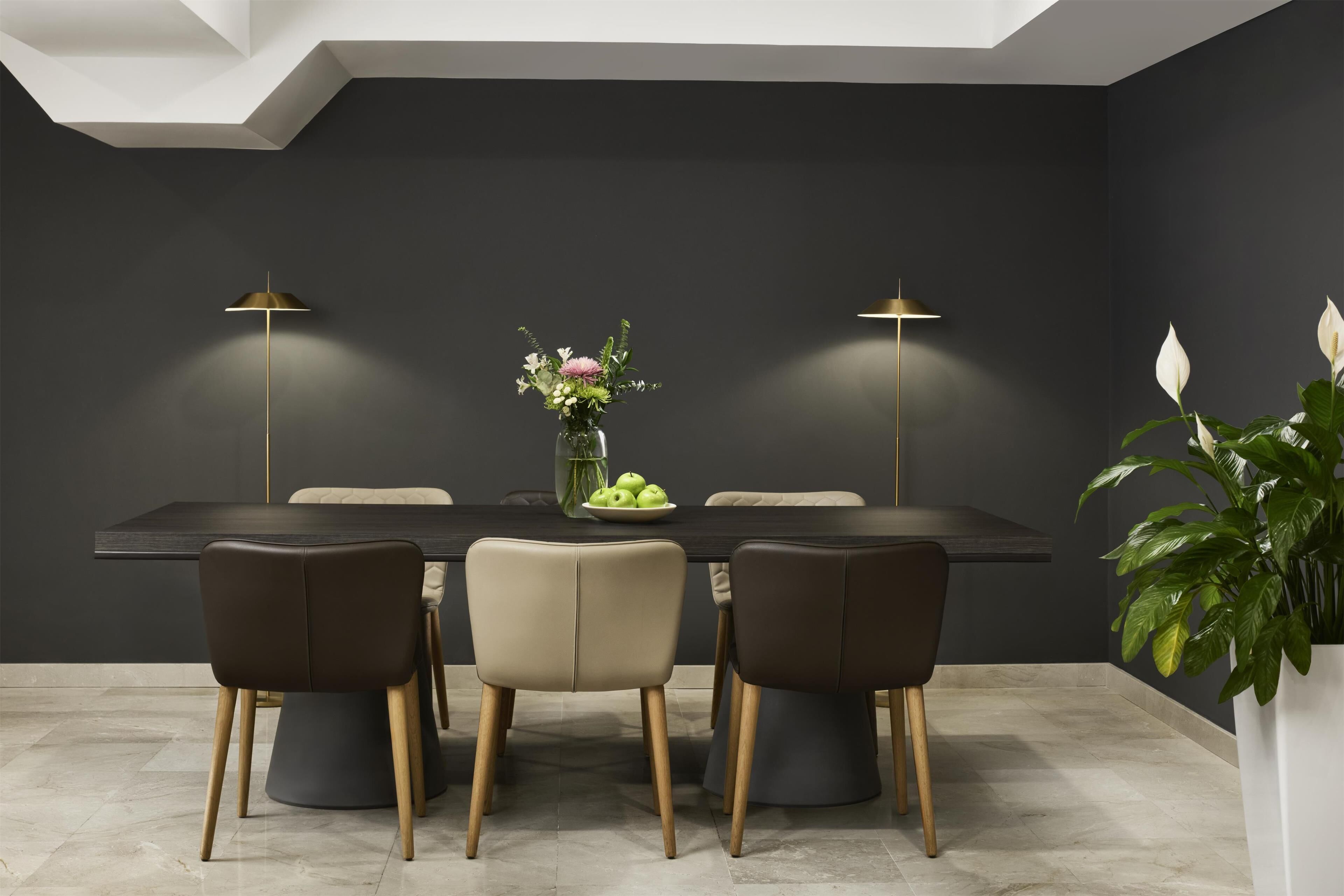 Un espace de salle à manger moderne avec un mur foncé, une table ronde et quatre chaises. On y trouve deux lampadaires, un vase avec des fleurs et une plante en pot sur la table.