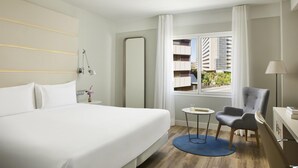 A hotel room with a large bed, a small table, a chair, a lamp, and a view of the city through a window. - NH Madrid Balboa (Madrid)
